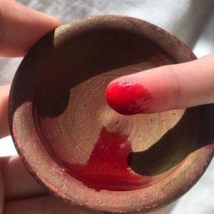 May include: A small, round, terracotta-coloured bowl with a gold rim, filled with a vibrant red substance. A finger is dipped into the red liquid, showing the texture and colour. The bowl is held in a hand.