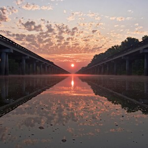 May include: A bridge spanning a body of water with a sunrise in the background. The bridge is constructed from concrete and features a series of arches. The water is still and reflects the sky and the bridge.