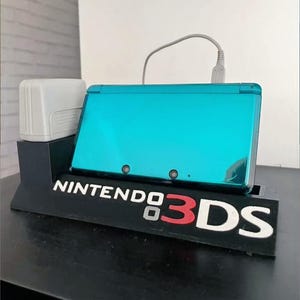 Può includere: Una console di gioco portatile Nintendo 3DS turchese su un supporto di ricarica nero. Il supporto presenta il logo Nintendo 3DS in bianco e rosso. È presente anche un adattatore di ricarica bianco.