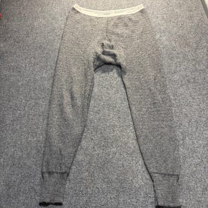 May include: A pair of heather gray thermal long underwear pants featuring a textured knit fabric, a light cream-colored elastic waistband, and fitted ribbed cuffs at the ankles. The garment has a full-length design with a tapered leg cut.