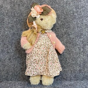 May include: A vintage teddy bear doll wearing a pink sweater and floral dress. The bear has a matching floral hat and is holding a smaller teddy bear. The doll has a classic, nostalgic design.