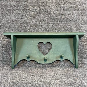 May include: A forest green wooden wall shelf featuring a flat top surface and a vertical back panel with a central heart-shaped cutout. The shelf includes three small, rounded wooden pegs extending from the front panel for hanging items.