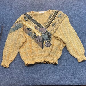 May include: A yellow long-sleeve sweater with a V-neckline and a textured knit pattern. The front features a decorative panel with dark green and gold fabric accents, embroidered floral motifs in shades of purple and blue, and ribbed cuffs.