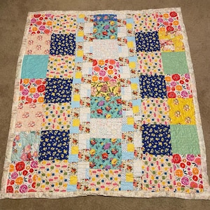 May include: A colorful patchwork quilt with a floral theme. The quilt features squares of various patterns, including floral prints, solid colors, and patterned designs. The color palette includes red, blue, yellow, and green. The quilt is bordered with a white fabric.