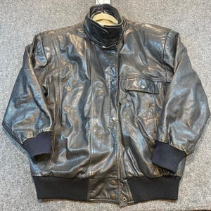 May include: A black leather jacket with a high collar and a front button closure. The jacket has two front pockets and ribbed cuffs and hem. The jacket is hanging on a hanger.