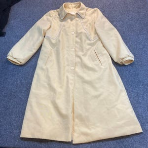 May include: A pale yellow long-sleeved coat featuring a pointed collar, a front button closure, and two side pockets. The garment includes white piping detail along the upper chest area and cuffs, with a structured silhouette and full-length hem.