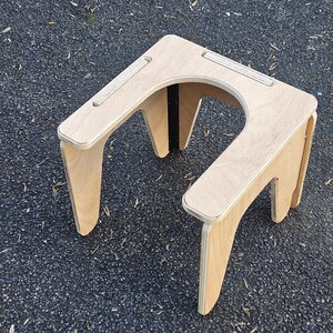 May include: A light-colored wooden stool with a U-shaped cutout. The stool has four angled legs and a flat top with two recessed grooves. The wood grain is visible, and the stool appears to be made of plywood.