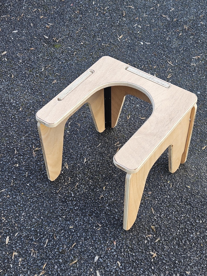 May include: A light-colored wooden stool with a U-shaped cutout. The stool has four angled legs and a flat top with two recessed grooves. The wood grain is visible, and the stool appears to be made of plywood.