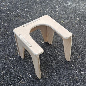 May include: A light-colored wooden stool with a square top and curved cutout. The stool has four angled legs and two parallel grooves on the top surface. The stool is made of plywood and has a natural wood grain finish.