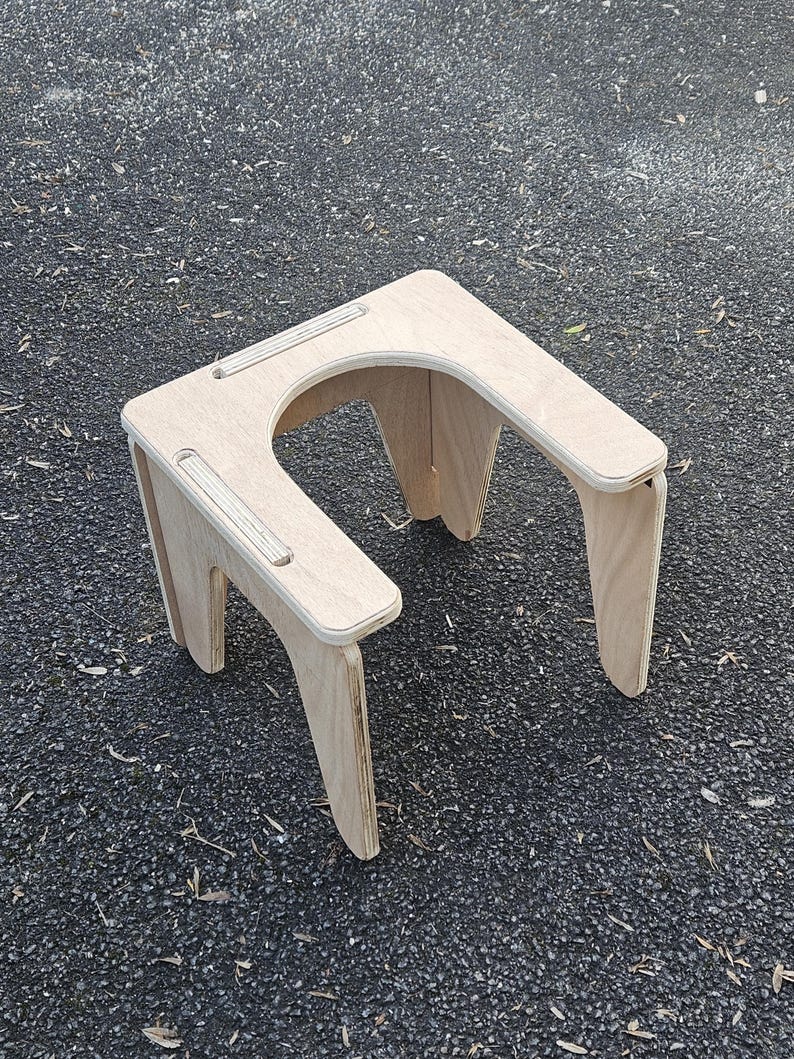 May include: A light-colored wooden stool with a square top and curved cutout. The stool has four angled legs and two parallel grooves on the top surface. The stool is made of plywood and has a natural wood grain finish.