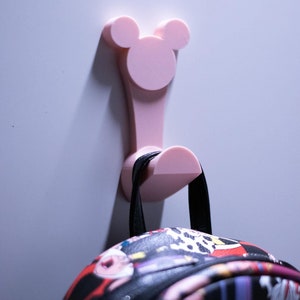 Pode incluir: Um gancho de plástico rosa em forma de Mickey Mouse montado em uma parede branca com uma mochila estampada em preto e vermelho pendurada.