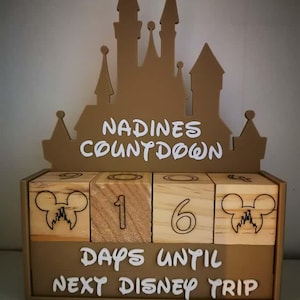 Pode incluir: Um calendário de contagem regressiva de madeira com uma silhueta de castelo marrom. O calendário exibe "Nadine's Countdown" e "Days Until Next Disney Trip" em letras brancas. Os blocos de madeira mostram o número "16" com silhuetas do Mickey Mouse.