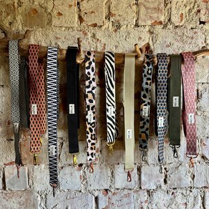 Könnte beinhalten: Eine Sammlung gemusterter Taschenriemen, die an einem Ast vor einer Backsteinmauer hängen. Die Riemen weisen verschiedene Designs auf, darunter geometrische Muster, Tiermuster und unifarbene Designs. Jeder Riemen hat Metallbeschläge.