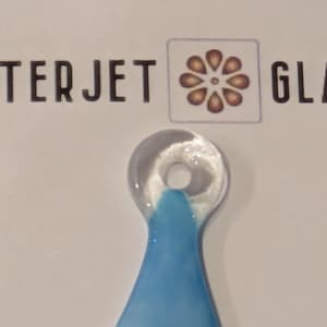 Op de afbeelding: Een heldere glazen hanger in de vorm van een druppel, met een blauwe gradiënt aan de onderkant. De tekst "WATERJET GLASS" staat boven de hanger, met een bloemenlogo. De hanger heeft een klein gaatje aan de bovenkant.