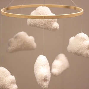 May include: A white, fluffy cloud mobile with six clouds hanging from a wooden ring. The clouds are made of yarn and are different sizes and shapes.
