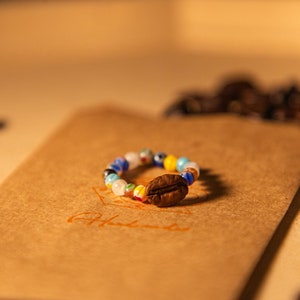 May include: A colorful beaded ring with a coffee bean charm on a brown paper envelope surrounded by coffee beans.