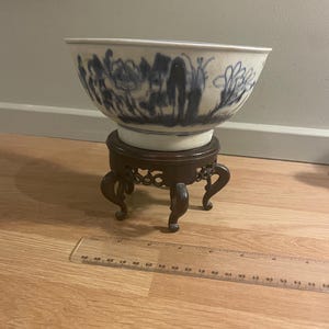 May include: A white and blue ceramic bowl with a floral design, on a dark brown wooden stand with curved legs. The bowl is about 25 cm in diameter. A decorative piece for the home.