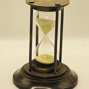 May include: An hourglass featuring a dark metallic frame with a circular base and top plate. The clear glass bulbs contain light-coloured sand, with a portion of the sand currently resting in the bottom bulb, held by four vertical metal support rods.