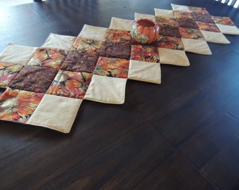 May include: A beige and brown quilted table runner with a floral pattern. The runner is made of fabric squares sewn together in a diamond pattern. A small ceramic pumpkin is sitting on the runner.
