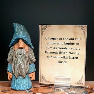 May include: A hand-carved wooden figurine of a bearded figure wearing a blue pointed hat and robe, standing next to a rectangular card with the text: A keeper of the old rain songs who begins to hum as clouds gather. Farmers listen closely, but umbrellas listen closer.