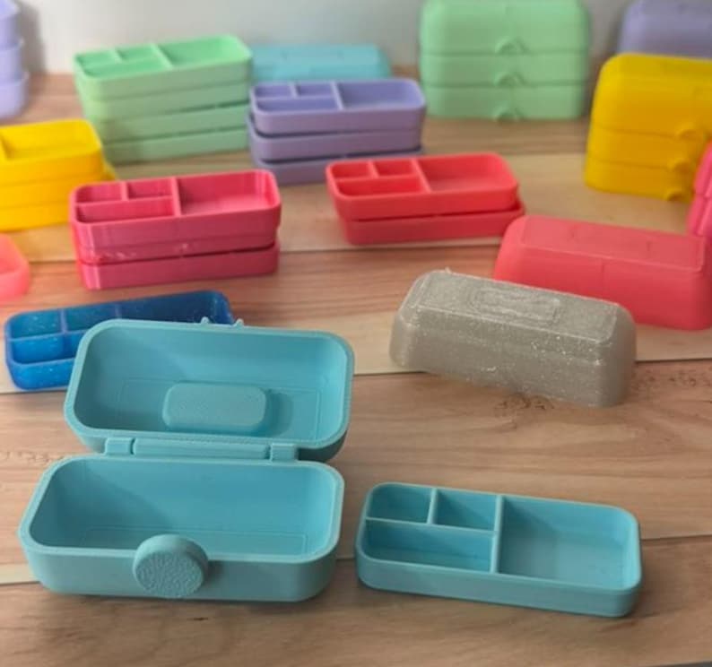 May include: A collection of colorful, small plastic organizers in various shades including blue, pink, green, yellow, and purple. Some are open, revealing compartments, while others are closed. The organizers are arranged on a wooden surface.