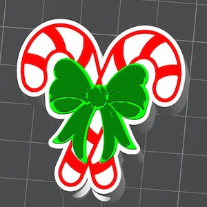 May include: A 3D-rendered Christmas decoration featuring two candy canes with red and white stripes, joined by a green bow. The design has a white outline and a shadow effect, suggesting a festive holiday theme.