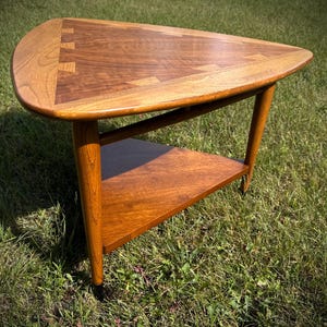 May include: A triangular wooden side table featuring a smooth, polished surface with contrasting light and dark wood grain patterns. The tabletop displays visible dovetail joinery, supported by three tapered legs and a lower triangular shelf.