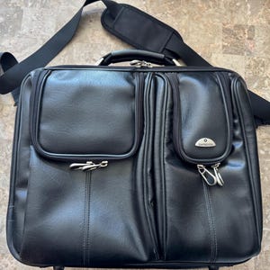 Puede incluir: Maletín Samsonite de cuero negro con múltiples compartimentos y herrajes plateados. Cuenta con un asa superior, una correa para el hombro desmontable y un bolsillo frontal con cierre de cremallera. Diseñado para transportar documentos y otros elementos esenciales.