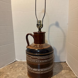 May include: A brown ceramic table lamp with a white and brown patterned design. The lamp has a pitcher-shaped base with a handle and a metal lampshade.