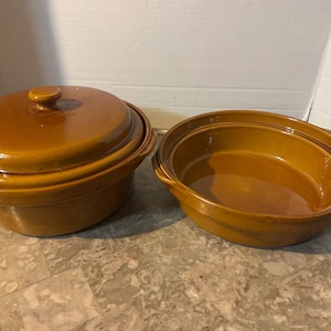 May include: Two brown ceramic casserole dishes. One dish has a lid with a round knob. The other dish is open. Both dishes have a glossy finish and are sitting on a patterned surface.
