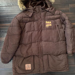 May include: A brown puffer jacket with a fur-lined hood. The jacket features a quilted design, two front pockets, and a logo patch. The jacket has the word "Maddox" in gold lettering on the chest.