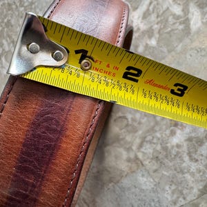May include: Close-up of a brown leather belt with a yellow tape measure showing a measurement of approximately 2 inches. The belt has a stitched edge and a worn appearance.