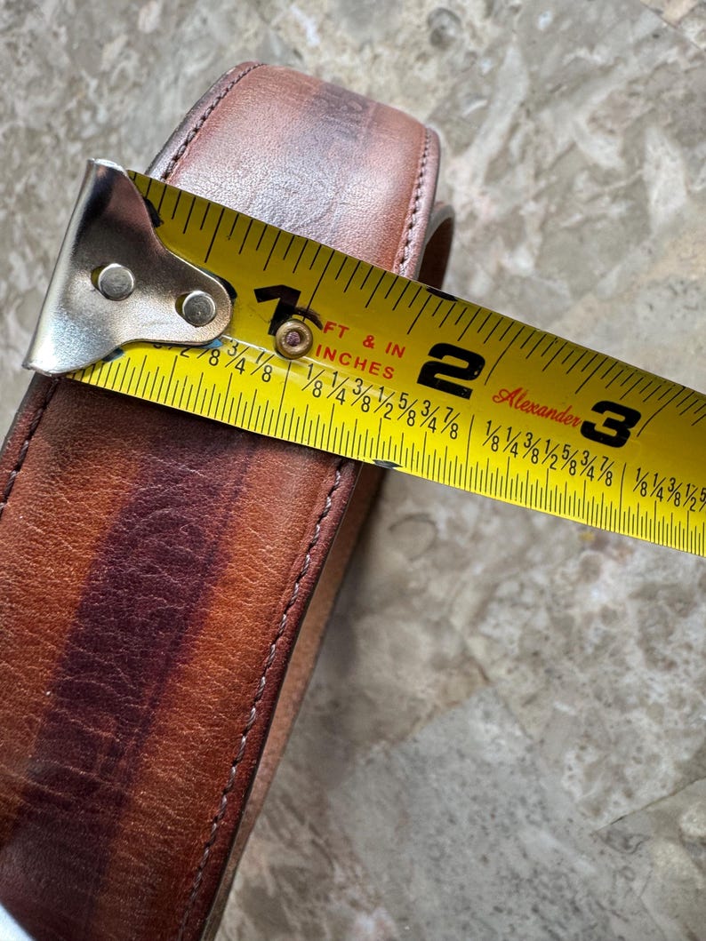 May include: Close-up of a brown leather belt with a yellow tape measure showing a measurement of approximately 2 inches. The belt has a stitched edge and a worn appearance.