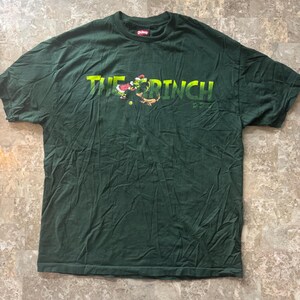 May include: Dark green t-shirt featuring the text "THE GRINCH" in yellow and green, with an illustration of the Grinch and Max. The shirt is made of a soft material.