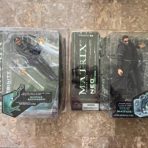 May include: Two action figures from The Matrix in original plastic packaging. The left package contains Trinity from Matrix Reloaded, and the right package contains Neo from Series One. Both boxes feature the Matrix logo and character names.