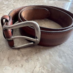 May include: A brown leather belt with a silver-colored buckle. The belt is coiled on a light-colored surface, showcasing its rich color and texture. The buckle is rectangular with a single prong.