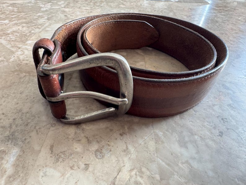 May include: A brown leather belt with a silver-colored buckle. The belt is coiled on a light-colored surface, showcasing its rich color and texture. The buckle is rectangular with a single prong.