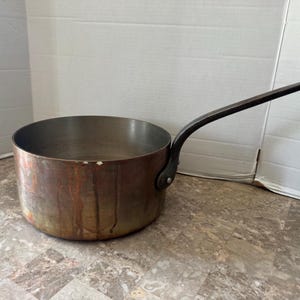 May include: A round copper saucepan with a long, dark metal handle attached by a single rivet. The exterior of the pan shows a weathered, tarnished patina with vertical streaks of discoloration, while the interior is a smooth, metallic silver color.