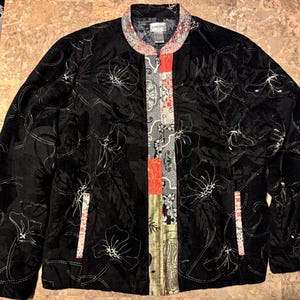 May include: Black jacket with floral embroidery and a colorful patterned panel down the front. The jacket has a stand-up collar and two pockets. The floral design is in a lighter color, creating a contrast against the black fabric.
