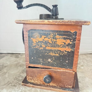 May include: Antique wooden coffee grinder with a black metal crank and drawer. The grinder has a rectangular wooden body with a faded black label that reads "E-Pound Coffee". The drawer has a black knob.