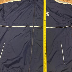 Op de afbeelding: Marineblauwe jas met capuchon, een ritssluiting over de hele lengte, witte en lichtblauwe strepen en een klein logo op de borst. De jas is ongeveer 76 cm lang, gemeten met een gele rolmaat.