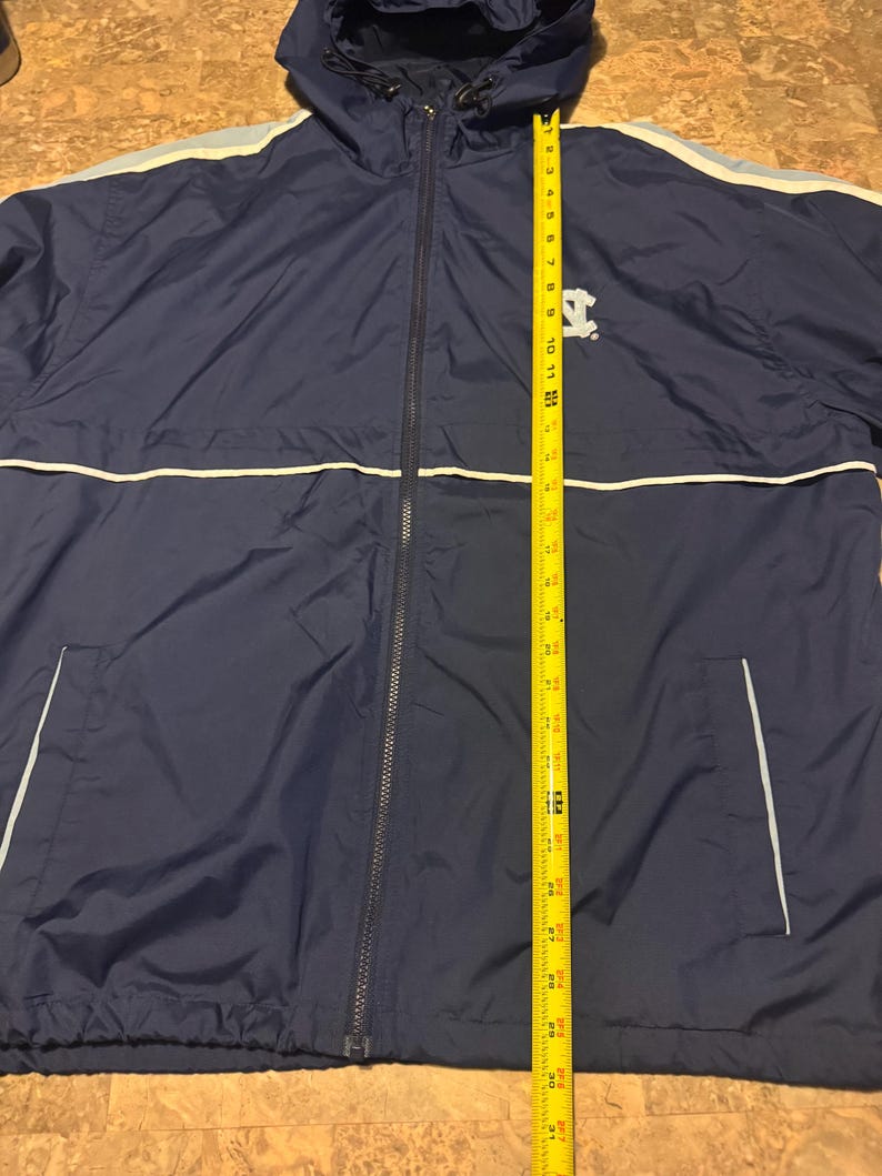 Op de afbeelding: Marineblauwe jas met capuchon, een ritssluiting over de hele lengte, witte en lichtblauwe strepen en een klein logo op de borst. De jas is ongeveer 76 cm lang, gemeten met een gele rolmaat.