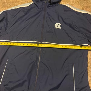 Op de afbeelding: Marineblauwe jas met capuchon, volledige ritssluiting en witte en lichtblauwe strepen op de mouwen. De jas heeft het logo van de University of North Carolina. De jas is ongeveer 61 cm lang.