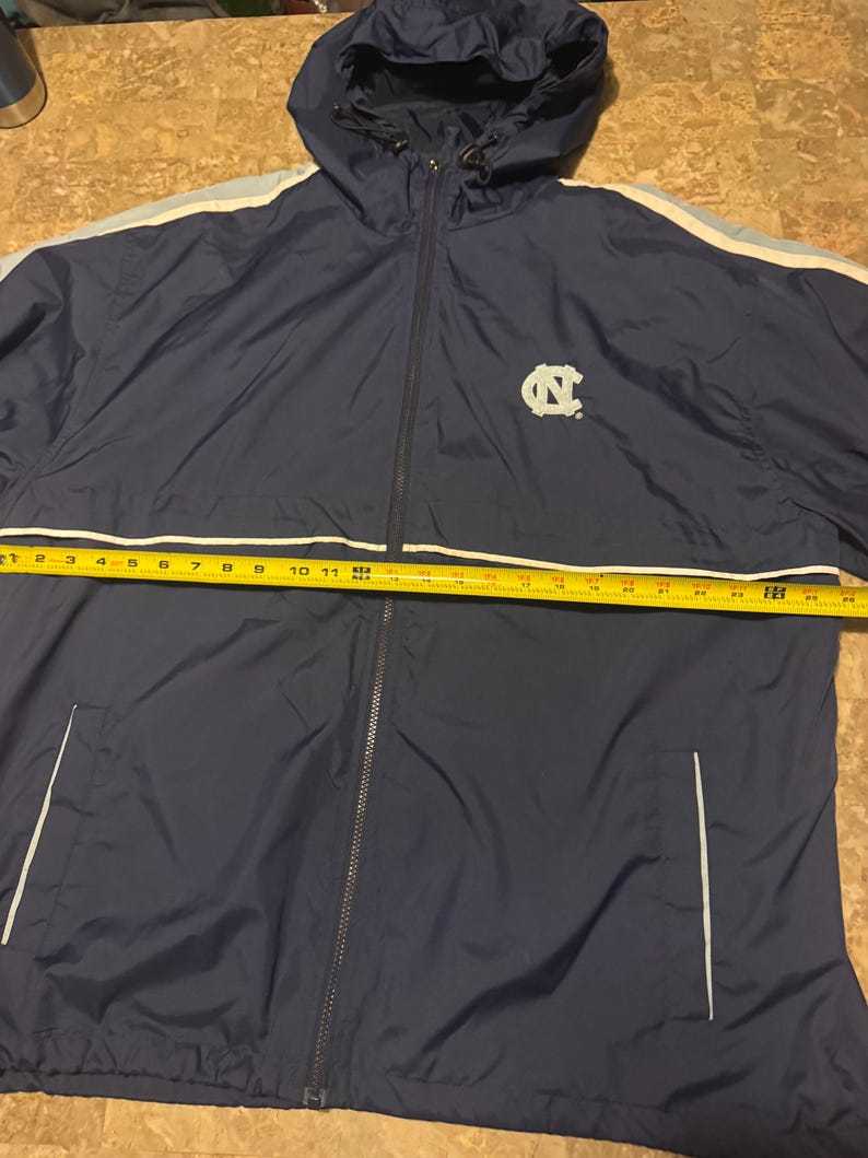 Op de afbeelding: Marineblauwe jas met capuchon, volledige ritssluiting en witte en lichtblauwe strepen op de mouwen. De jas heeft het logo van de University of North Carolina. De jas is ongeveer 61 cm lang.