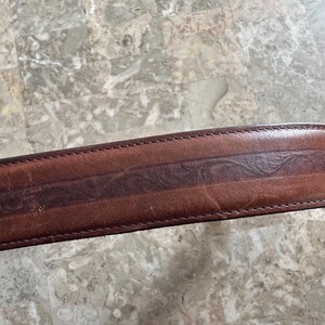 May include: Close-up of a brown leather belt with a decorative embossed design. The belt has a darker brown central stripe with a repeating pattern and is stitched along the edges.