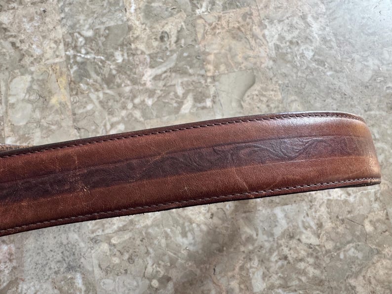 May include: Close-up of a brown leather belt with a decorative embossed design. The belt has a darker brown central stripe with a repeating pattern and is stitched along the edges.