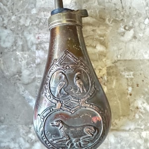 Könnte beinhalten: Antike Pulverflasche aus Metall in Tropfenform. Sie zeigt detaillierte Reliefs von Vögeln und einem Hund. Der obere Teil hat einen Messingausguss und einen Hebel. Das Metall hat ein angelaufenes, gealtertes Aussehen.