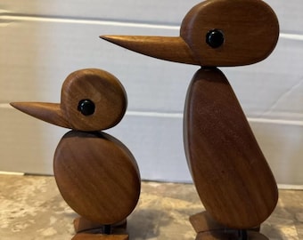 May include: Two wooden bird sculptures, one larger than the other, with black eyes. The birds are carved in a simple, modern style and have a natural wood finish.