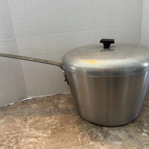 Op de afbeelding: Een zilverkleurige aluminium steelpan met een bijpassend deksel en een lange, platte metalen handgreep. Het deksel is voorzien van een kleine, donkerbruine, vierkante knop in het midden. De pan heeft een taps toelopende, cilindrische vorm.