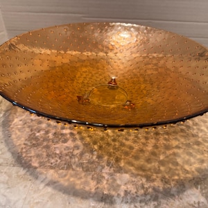 Może przedstawiać: Duża, bursztynowa szklana miska o teksturowanej powierzchni i centralnej, podwyższonej podstawie. Misa ma szeroki, płytki kształt i jest prawdopodobnie używana do celów dekoracyjnych lub serwowania. Obręcz jest ciemna, a miska rzuca cień.